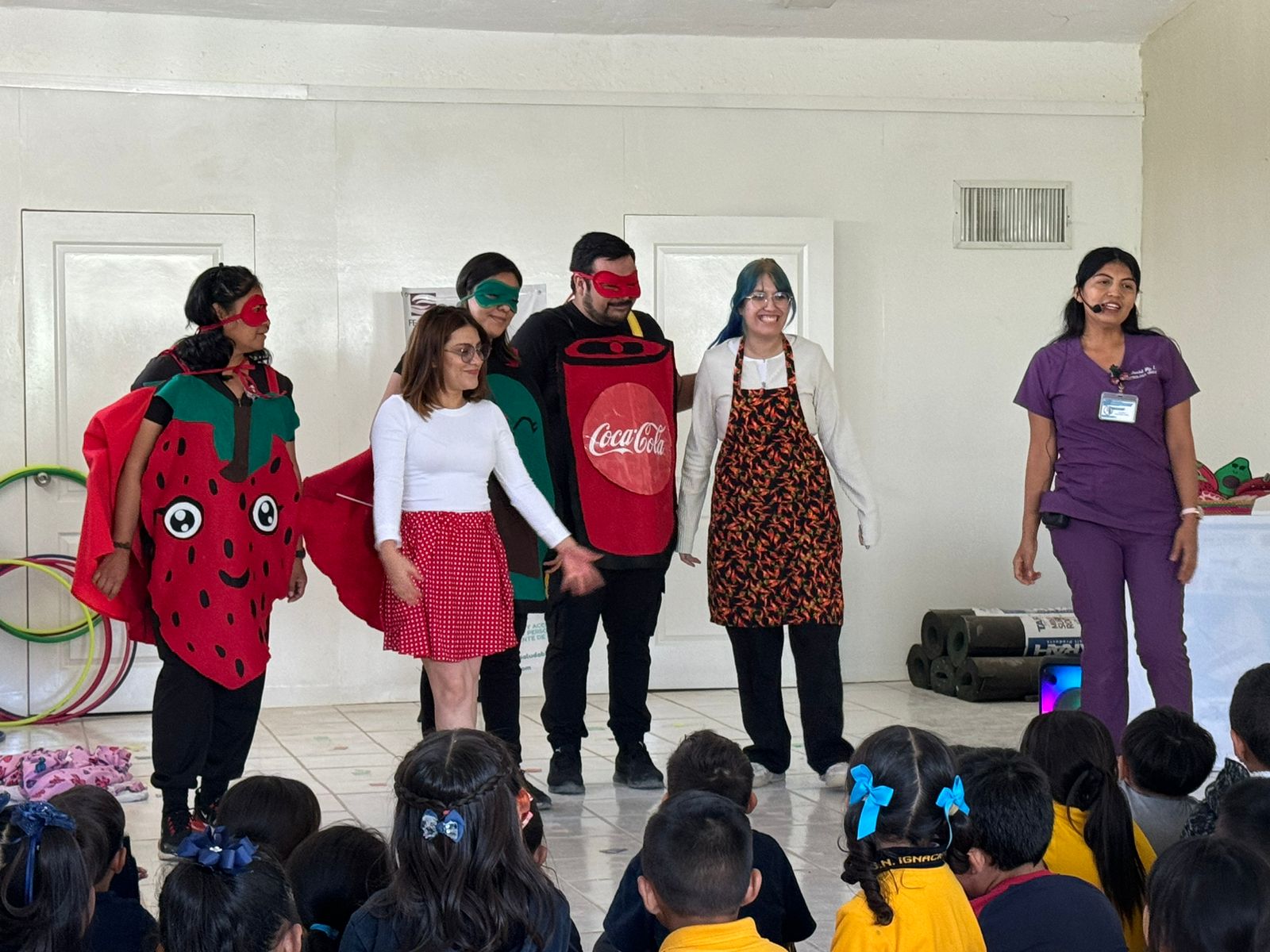 Grupo de niños y una profesora en un aula, con un pizarrón decorado y varias sillas dispuestas alrededor.