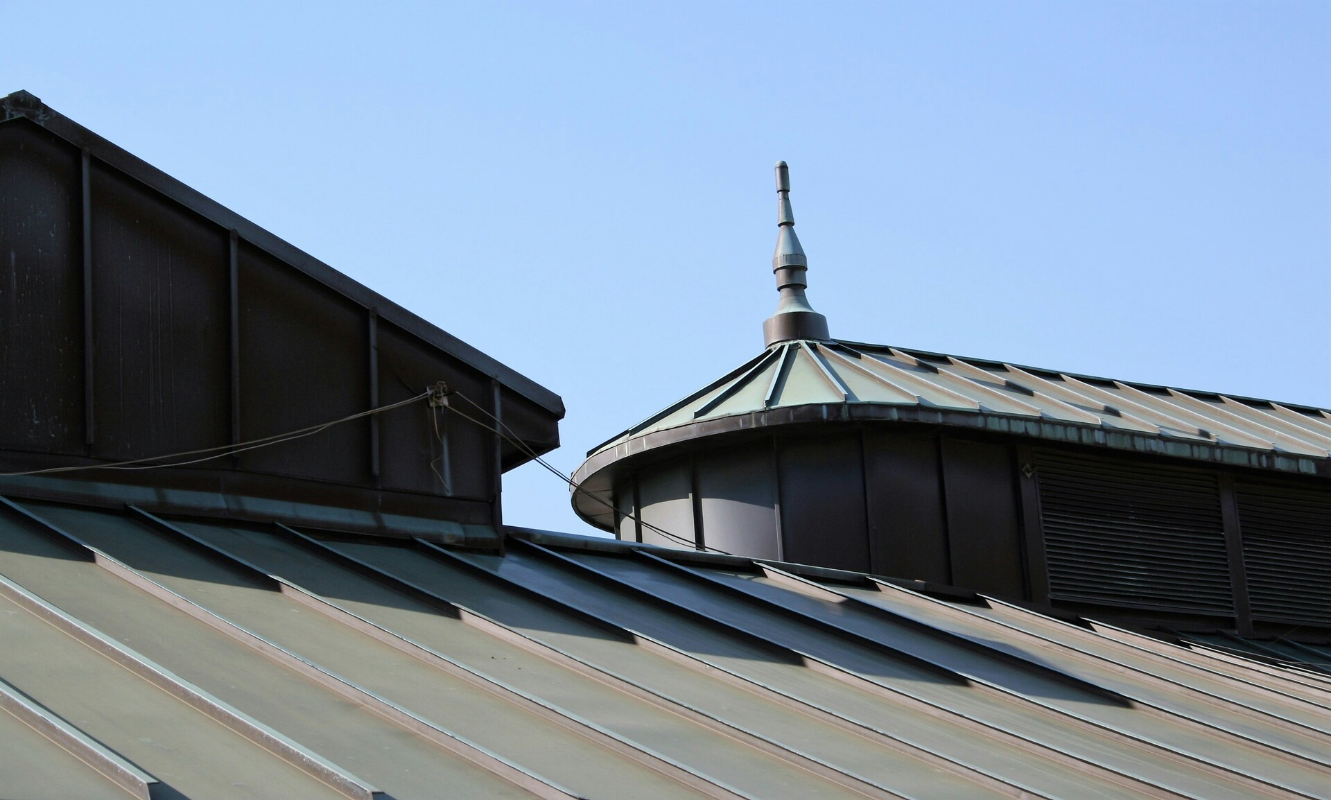 Metal building rooftop with green standing seam roof, dormer, vents, and decorative spire under blue sky