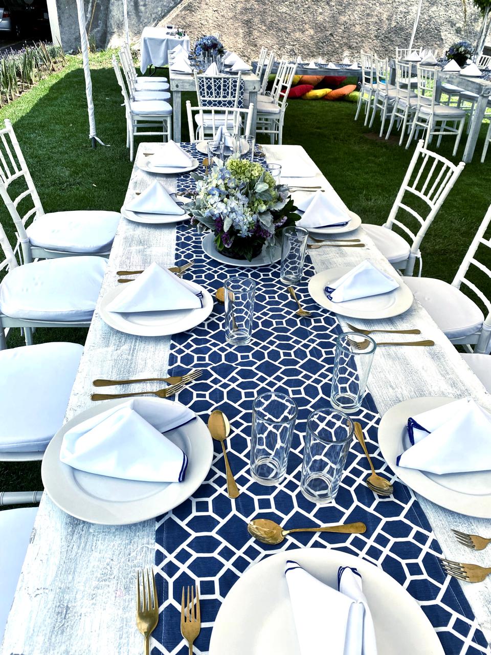 Mesa al aire libre decorada para evento, con mantel azul geométrico, cubiertos dorados, platos blancos y flores en el centro. Mesa al aire libre decorada para evento, con mantel azul geométrico, cubiertos dorados, platos blancos y flores en el centro.