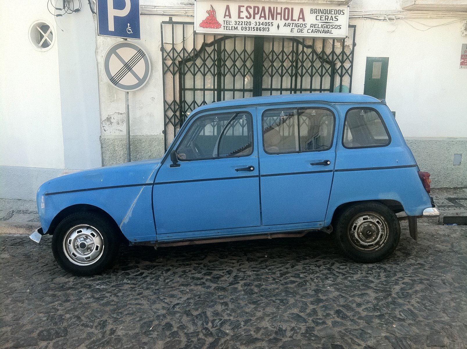 A blue car parked on a cobbled street with white walls, a metal gate and signs.