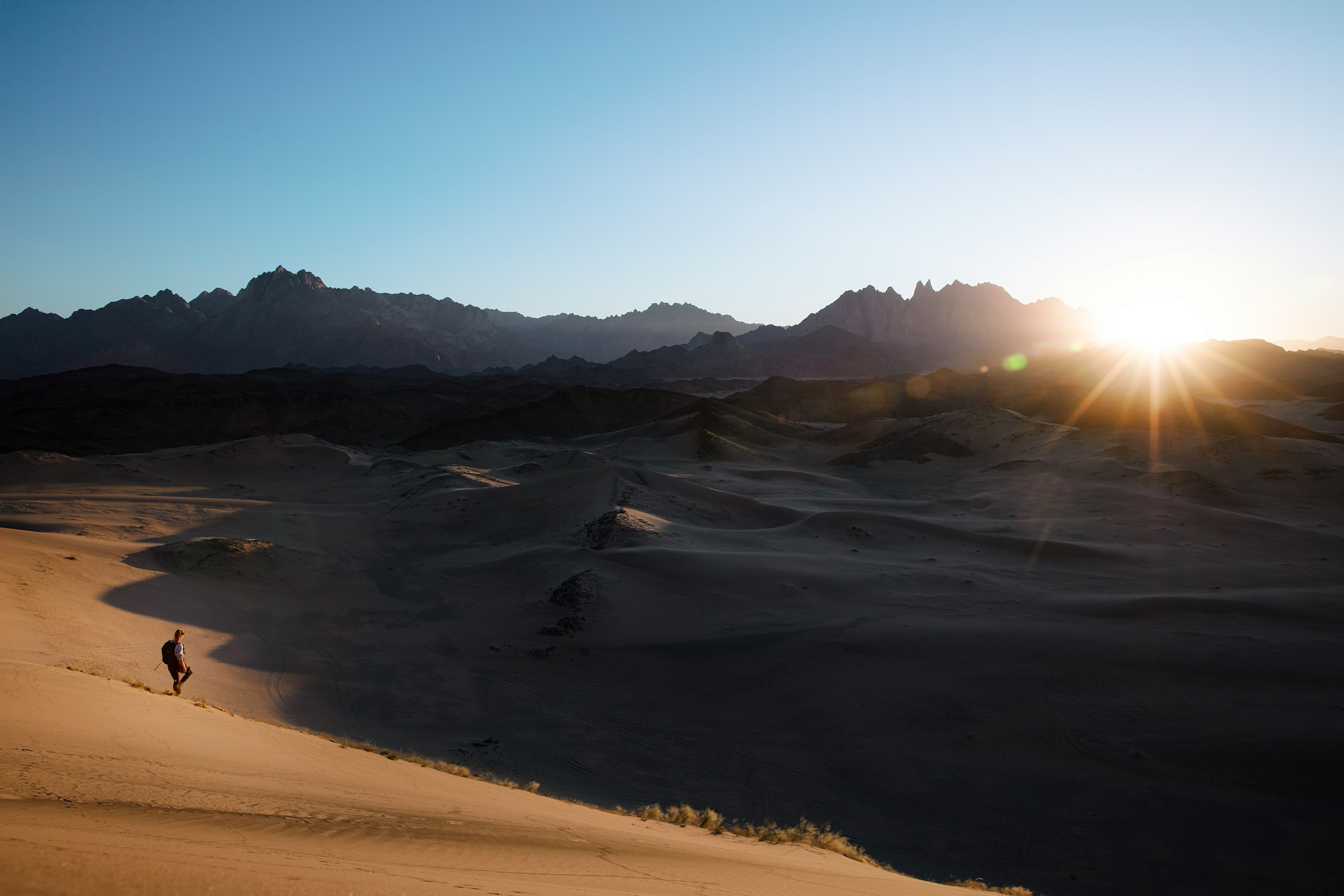 Amanecer en el desierto con montañas al fondo y una persona caminando por las dunas de arena.