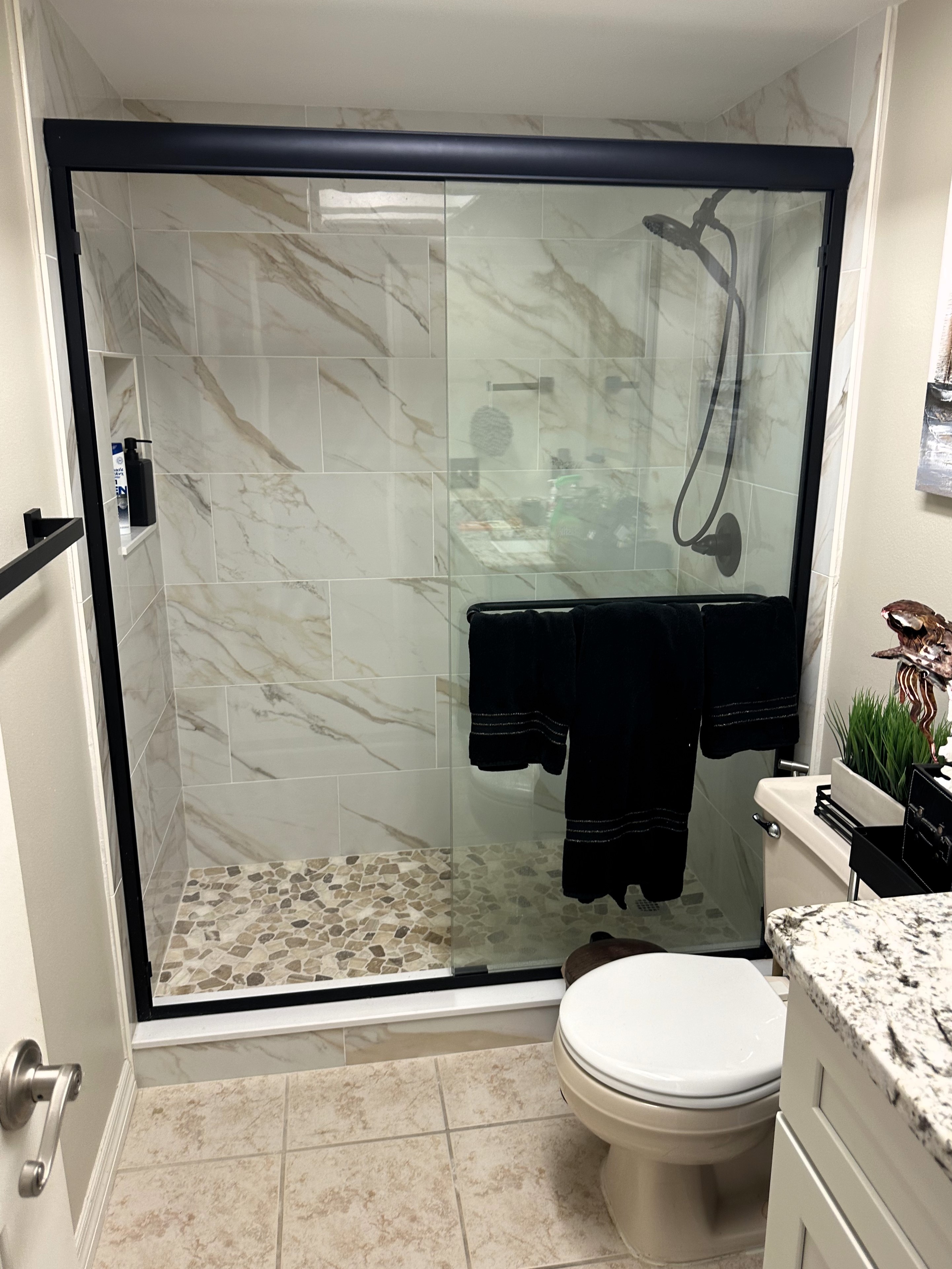 Lovely walk-in shower featuring matte black by-passing shower doors with flat stone-colored pebbles on the shower floor and 12x24 polished wall tiles.   