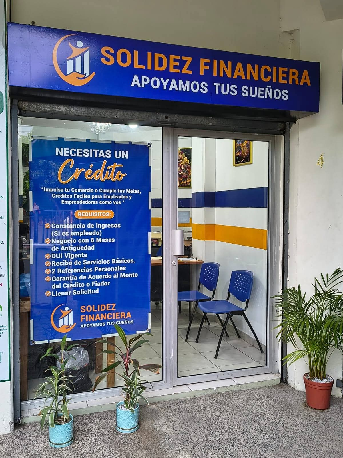 Fachada de Solidez Financiera con rótulo azul y anuncio de créditos, oficina de préstamos con puerta de vidrio y plantas