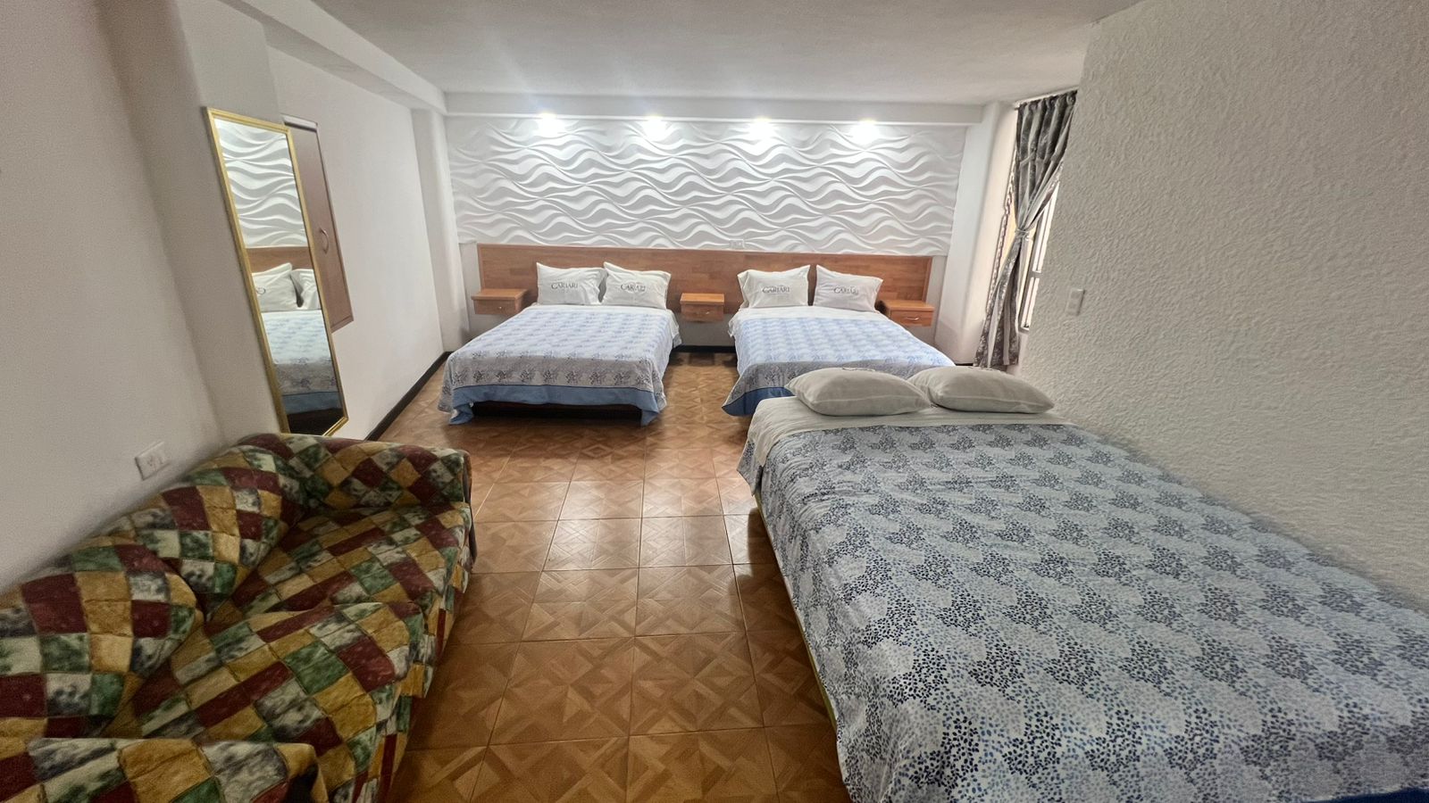 Habitación con tres camas, pared decorativa ondulada, espejo vertical y sofá con diseño de rombos.