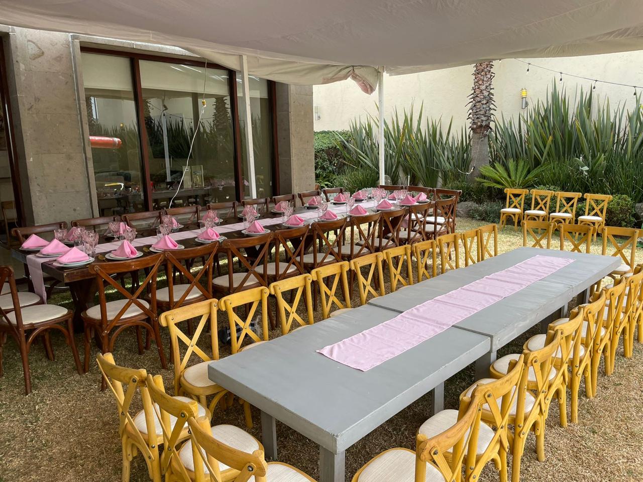 Mesa larga con sillas cruzadas de madera, decorada con servilletas rosas en un jardín exterior. Mesa larga con sillas cruzadas de madera, decorada con servilletas rosas en un jardín exterior.