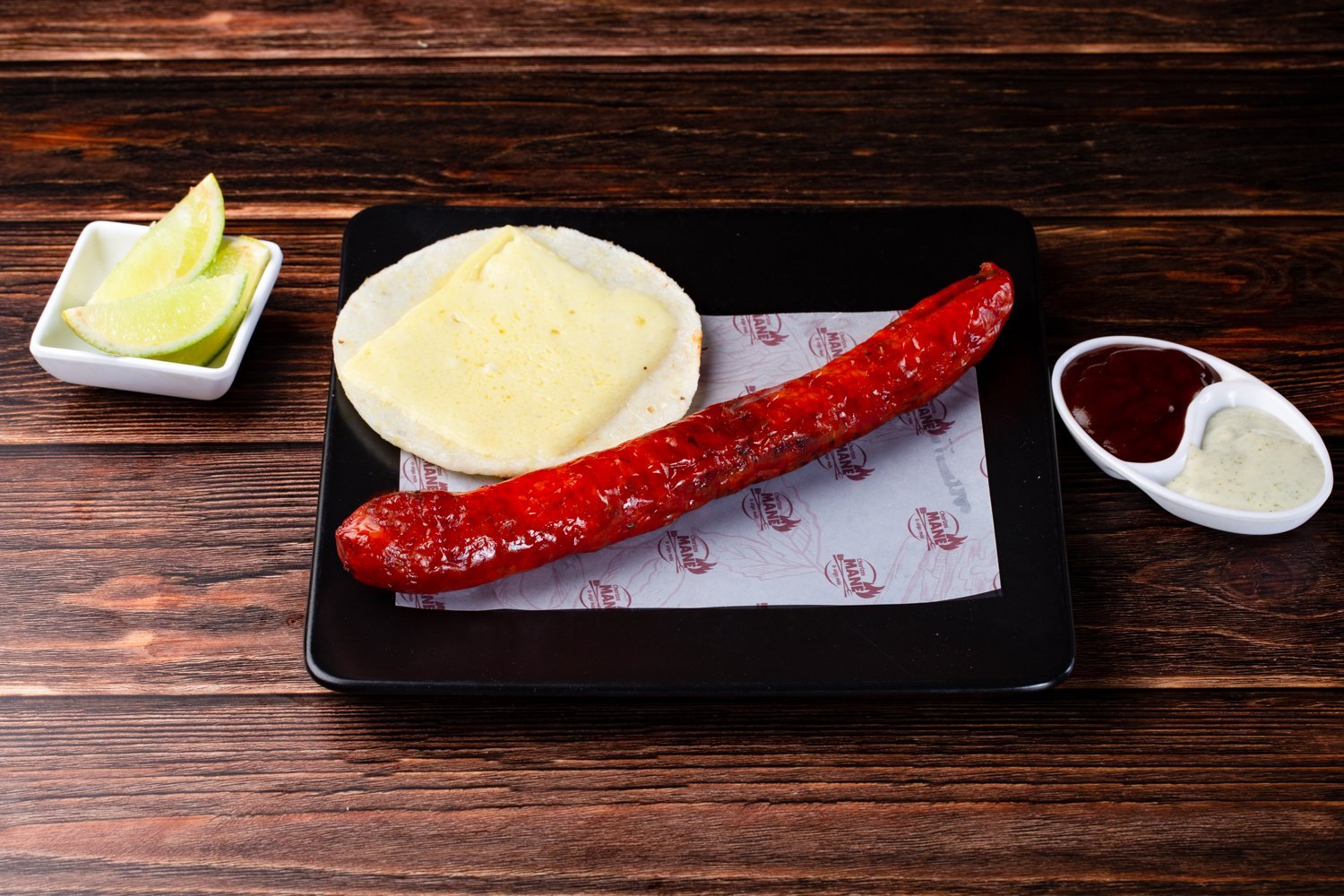 Chorizo asado con tortilla y queso sobre un plato, acompañado de salsas y rodajas de limón.