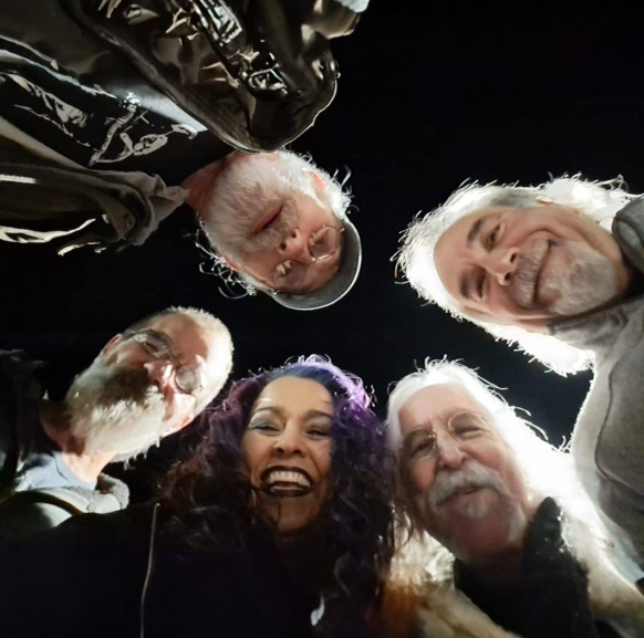 Group of five people smiling in a circular formation, taken from a low angle against a dark background.