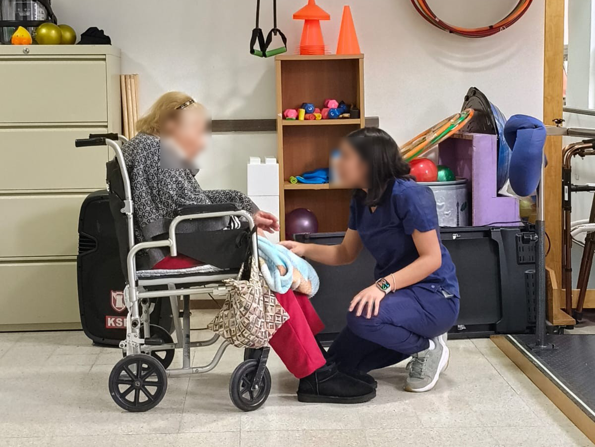 Elderly woman receiving caregiver assistance during a cognitive therapy activity with markers and grid worksheet