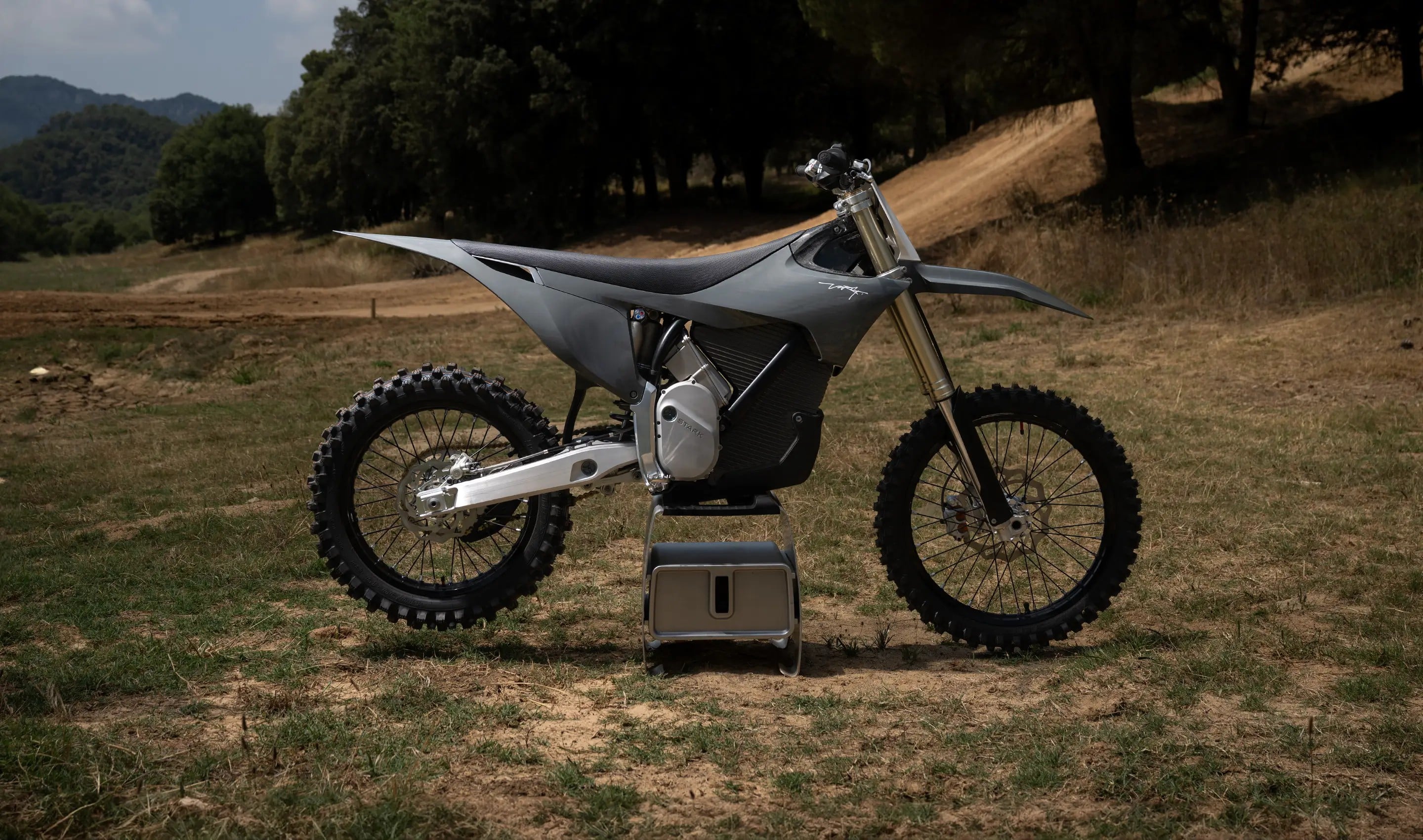 Motocicleta eléctrica de motocross gris en un sendero de tierra rodeado de árboles y montañas