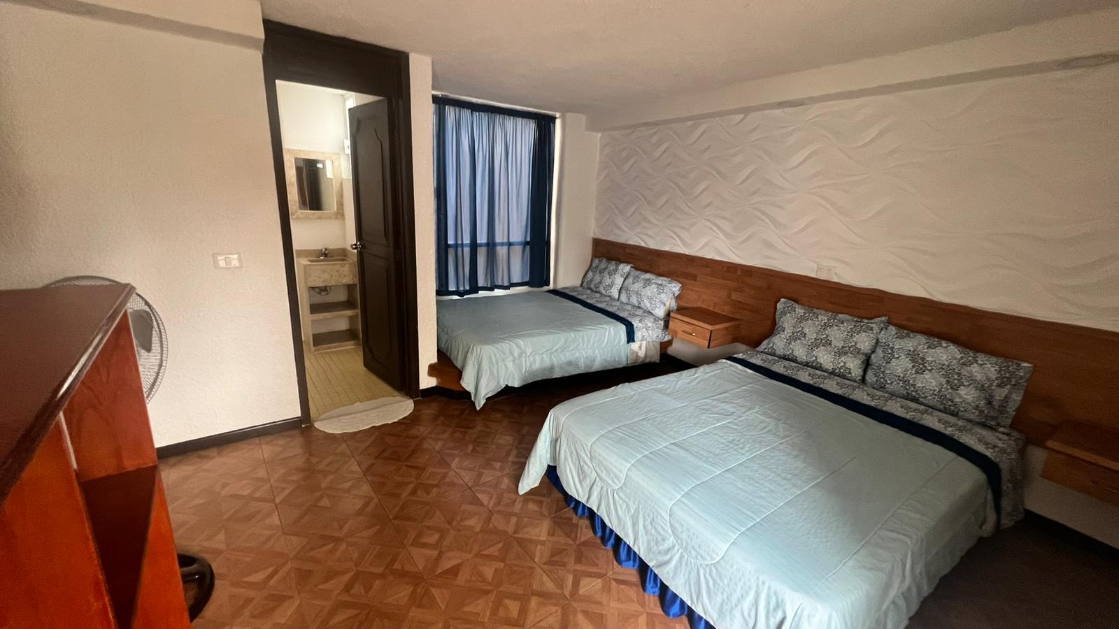 Habitación de hotel con dos camas matrimoniales, decoración en tonos azul y blanco, baño adjunto visible a través de una puerta abierta.