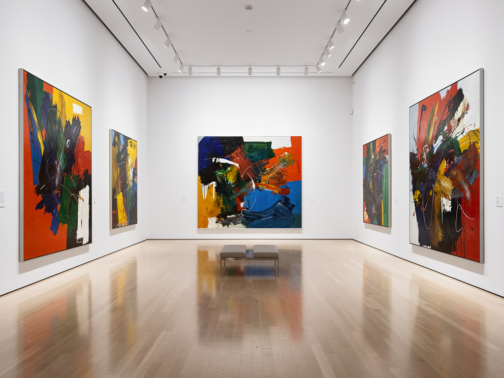Sala de galería de arte moderno con grandes pinturas abstractas coloridas, paredes blancas, iluminación de museo y suelo de madera pulido