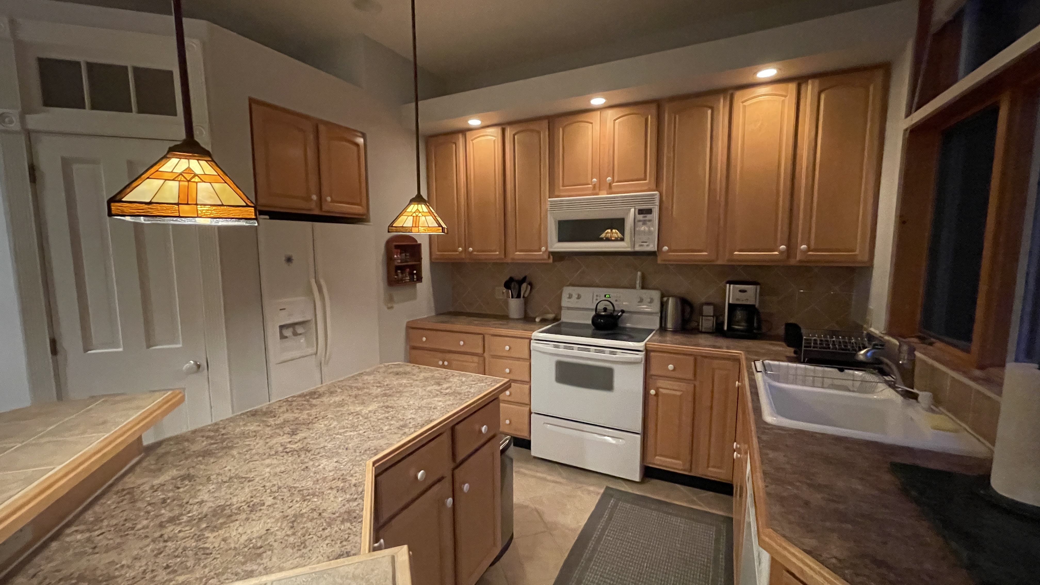 A warm, inviting kitchen with wooden cabinets, pendant lighting, white appliances, and a tiled countertop with a central island.