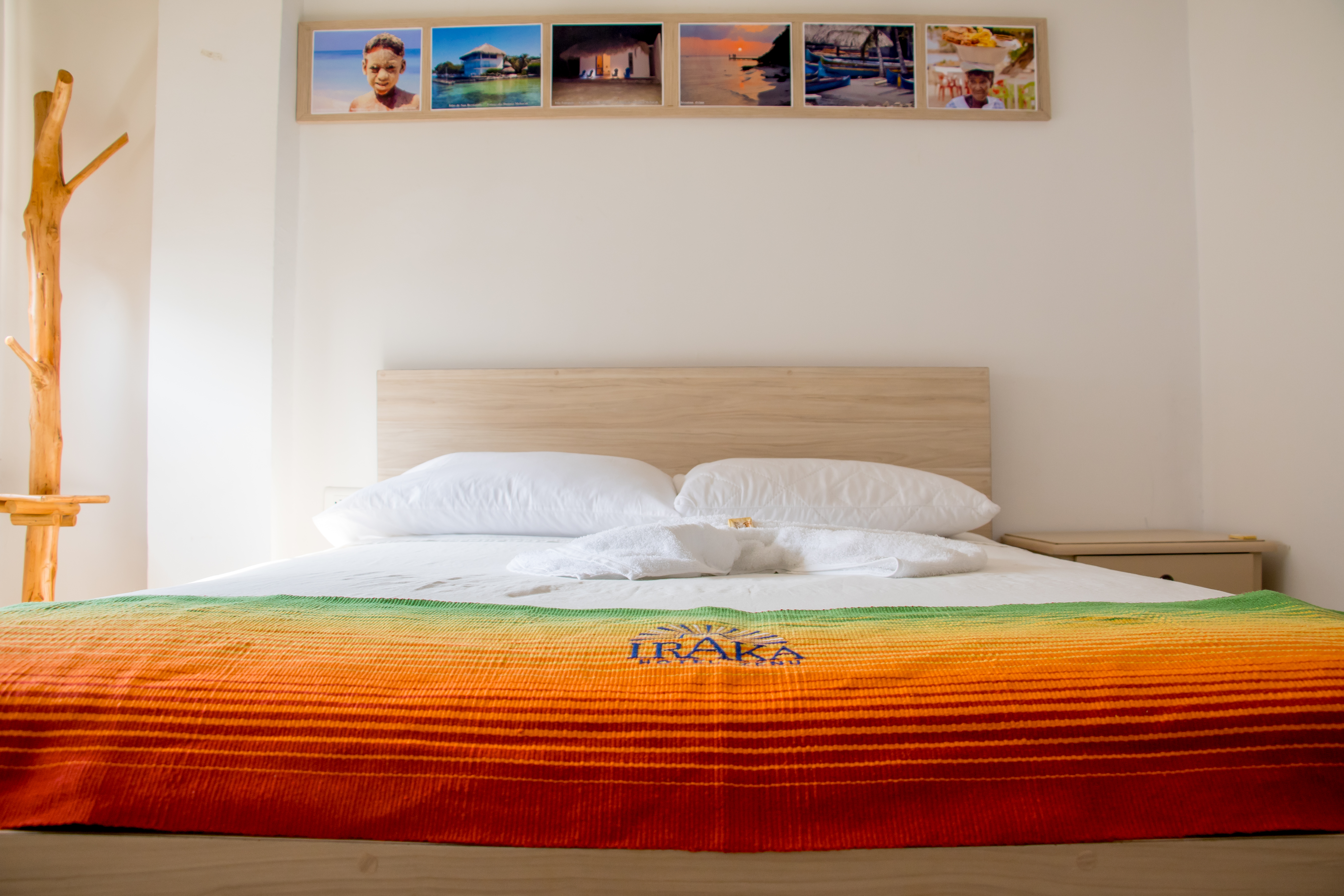 Cama con cabecera de madera, ropa de cama blanca, manta colorida con logo y cuadros decorativos en la pared.
