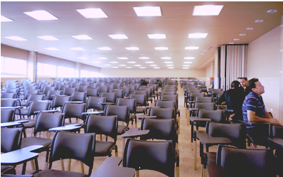 Aula vacía con filas de asientos y techo iluminado, vista desde el frente.