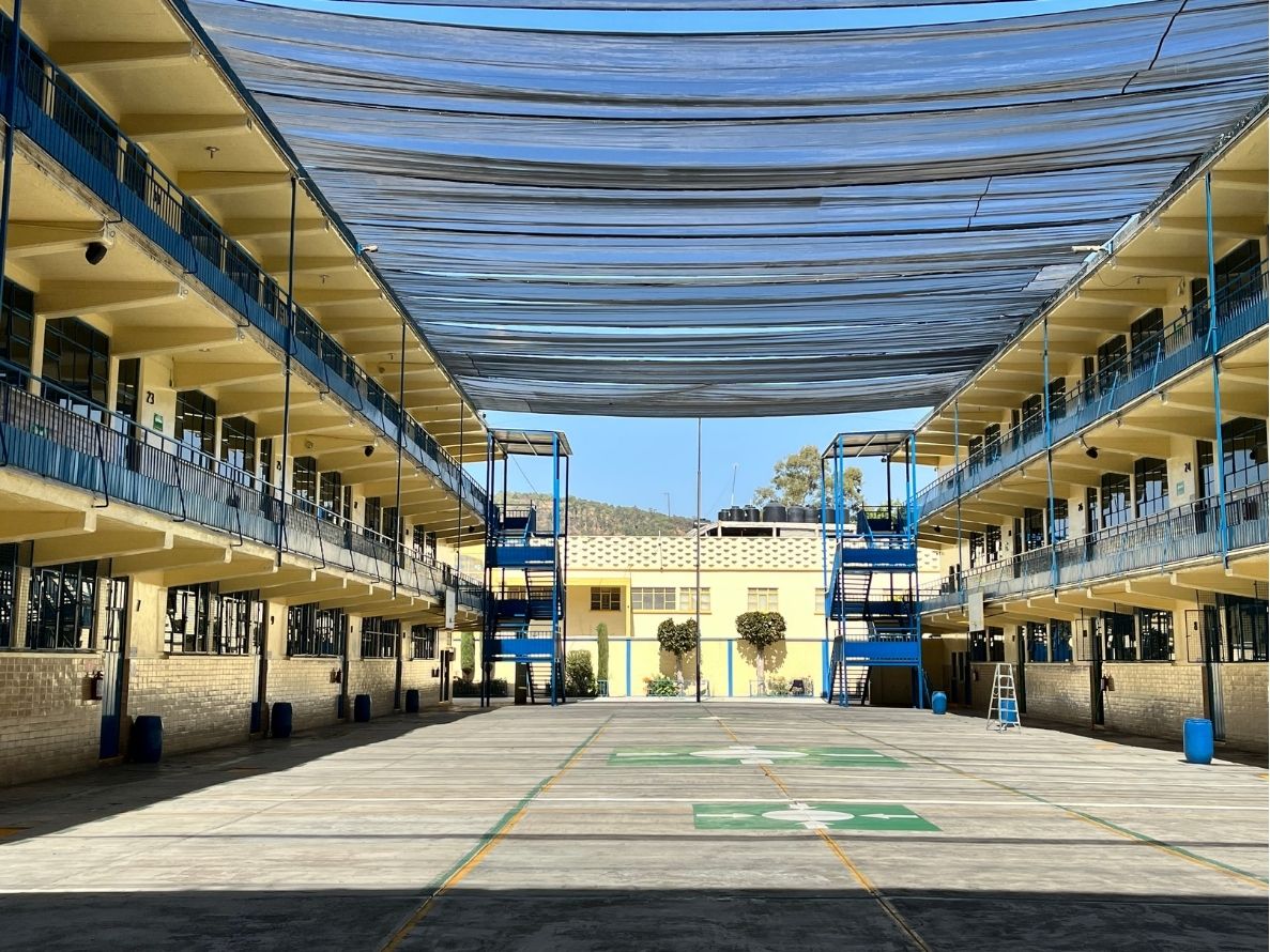 Patio escolar amplio con edificios de aulas, barandales azules y malla sombra en un día soleado