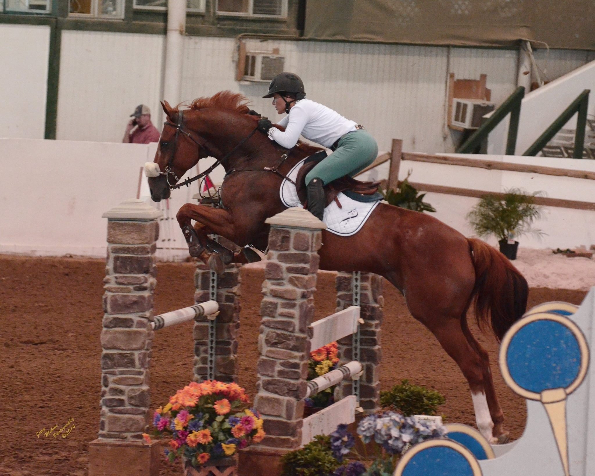Equestrian jumping competition with rider on chestnut horse leaping over stone obstacle in indoor arena.