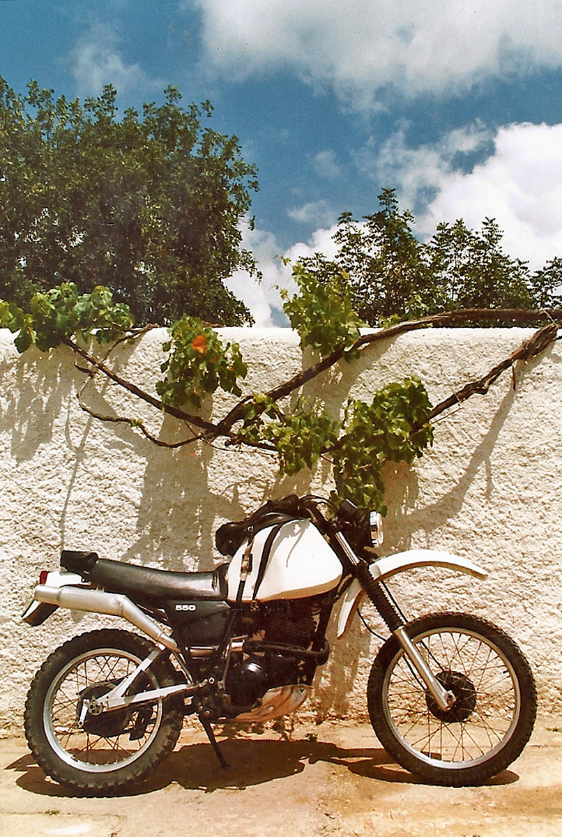 A white motorcycle with high fenders and knobby tires in front of a white wall.
