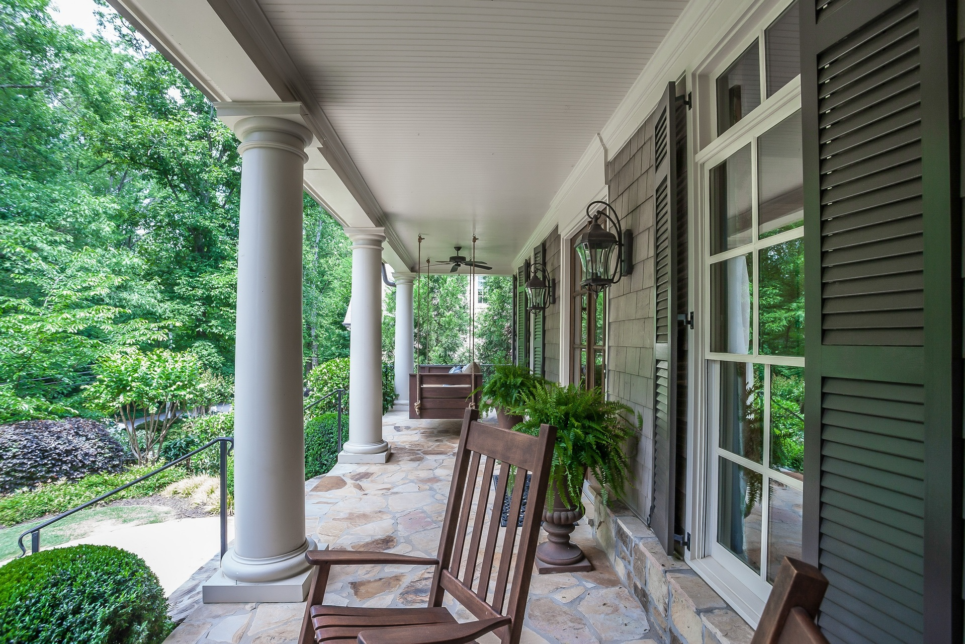 Charming porch with white columns, rocking chairs, hanging swing, and lush greenery.