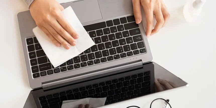 Persona limpiando teclado de portátil con un paño blanco, gafas en la mesa.