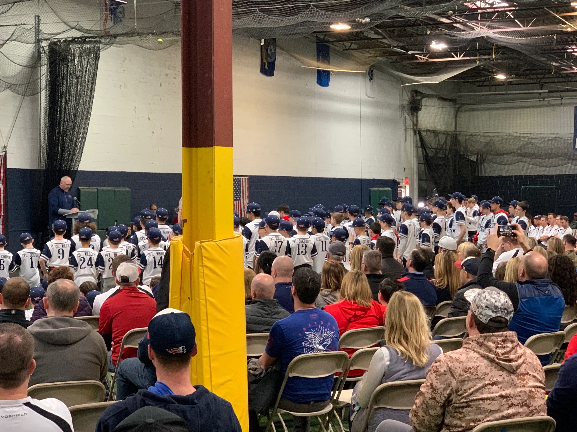 Indoor sports event with an audience seated in chairs watching a group of athletes in baseball uniforms listening to a speaker.