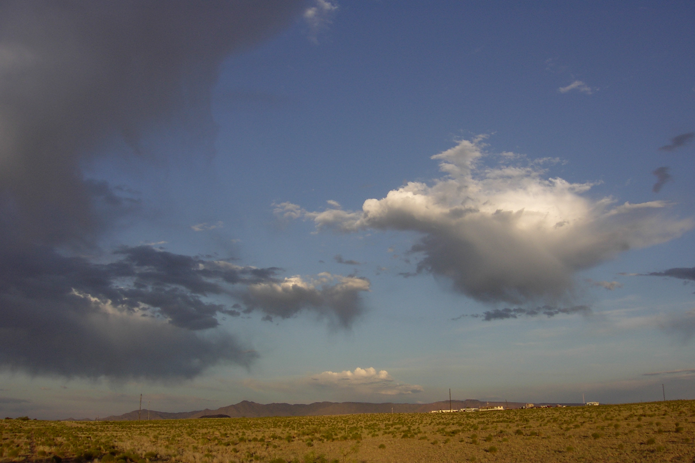 Desert landscape with a straight dirt road surrounded by dry bushes under a blue sky, leading towards distant mountains.