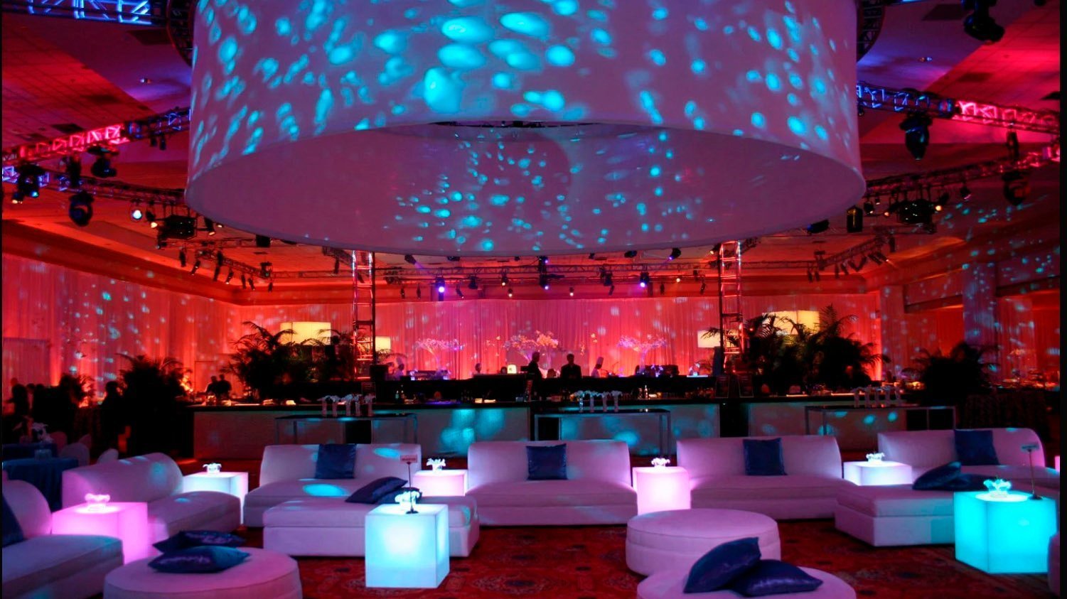 Evento en salón iluminado con luces azules y rosadas, sofás blancos y mesas brillantes, decoración moderna y acogedora.