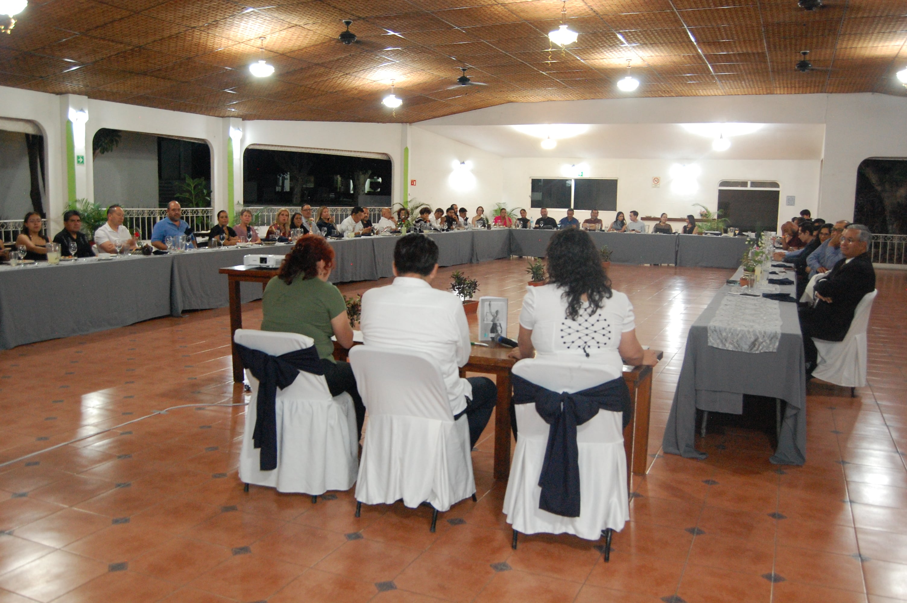 Personas sentadas alrededor de una gran mesa en una reunión formal en un salón iluminado.