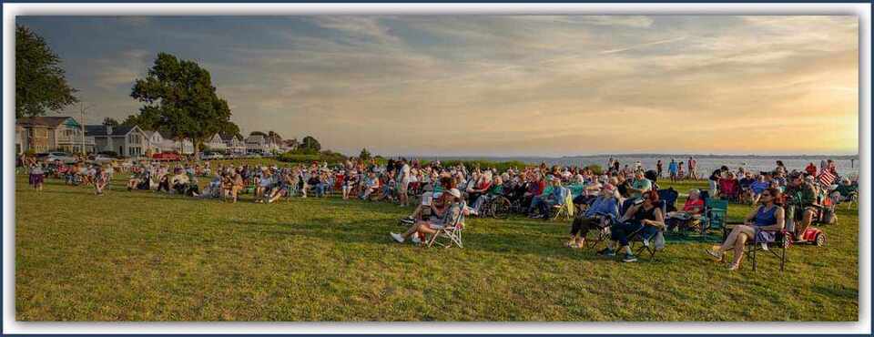 Crowd enjoying an outdoor event at sunset near a waterfront, surrounded by green grass and nearby houses.