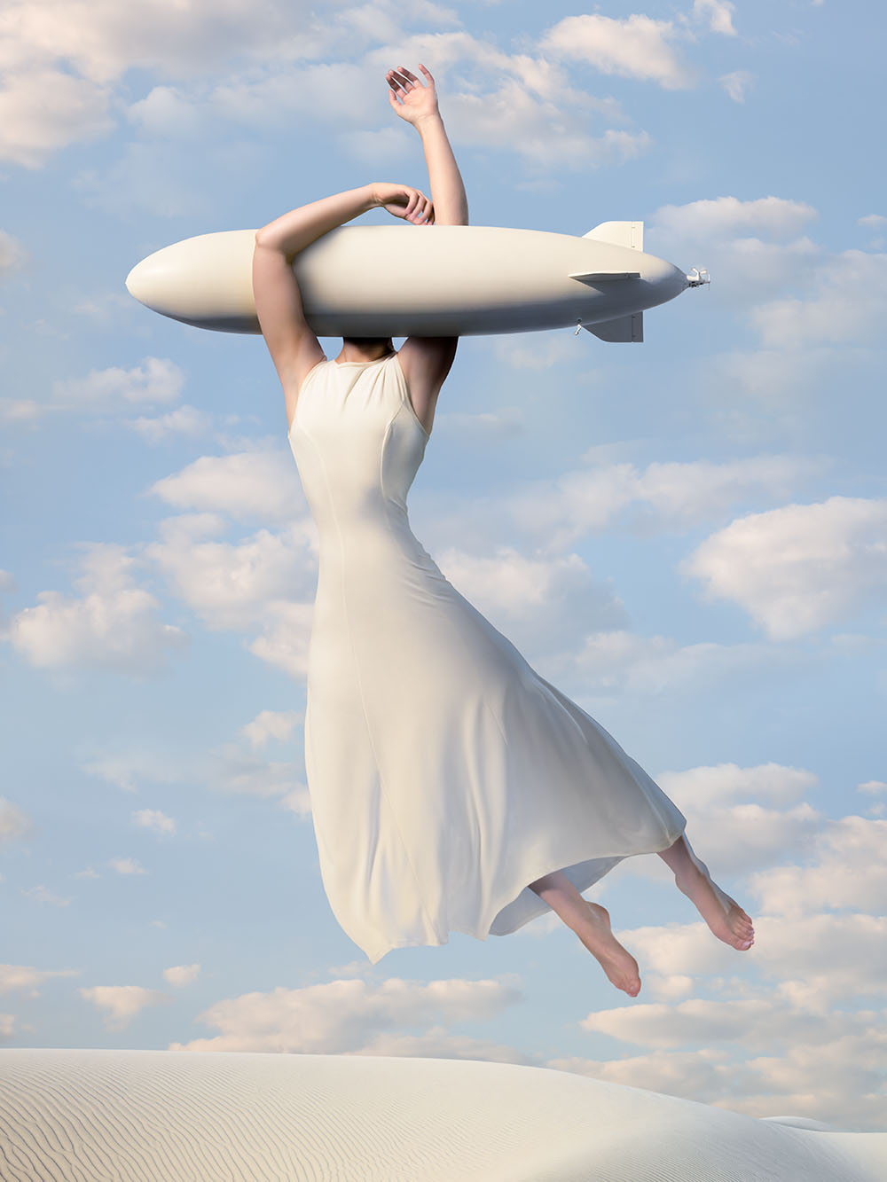 Dreamlike photo composite of a woman with head of a dirigible floating over dunes in cloud-filled sky.