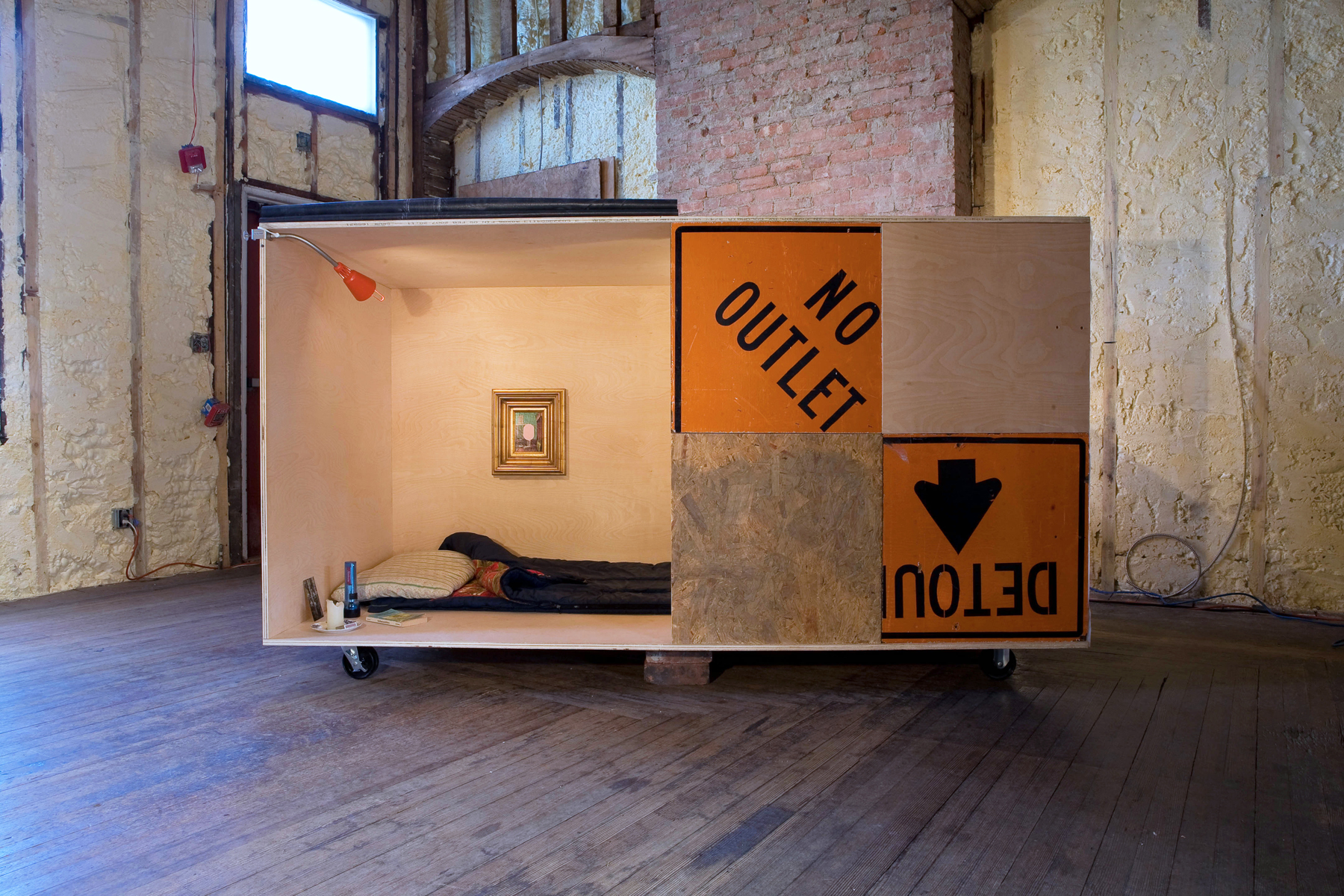 A box made from plywood and orange signs contains a bed and a painting.