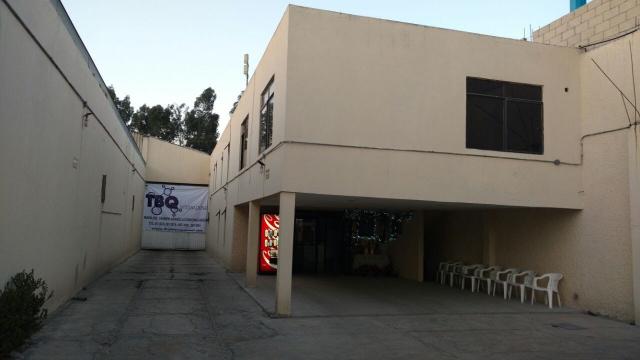 Edificio de dos pisos con sillas blancas en el exterior y una máquina expendedora en la entrada, rodeado de paredes altas.