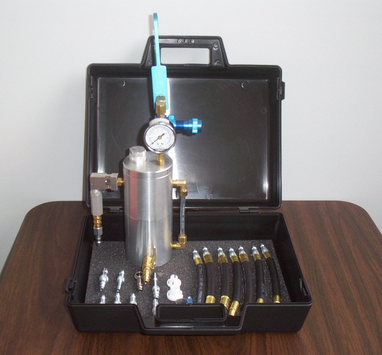 Portable pressure gauge kit with metal cylinder and hoses in a black carrying case on a wooden table.