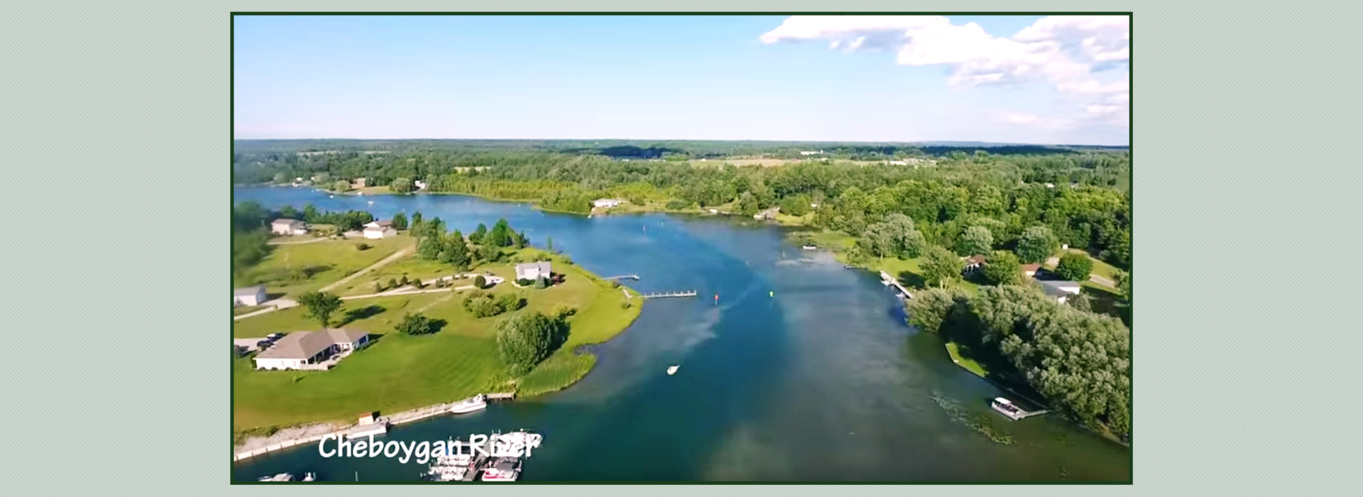Aerial view of Cheboygan River with surrounding green landscape and houses, clear blue sky above.