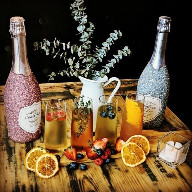 Elegant display of sparkling wine bottles with four cocktails garnished with berries and herbs, surrounded by sliced oranges and strawberries on a rustic wooden table.