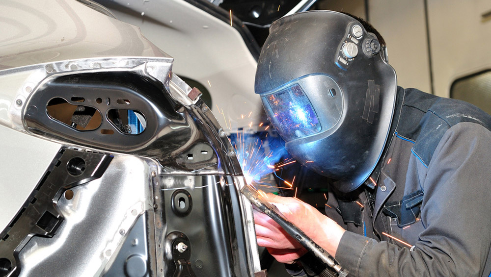 Soldador reparando la carrocería de un automóvil con máscara protectora y chispas en un taller mecánico