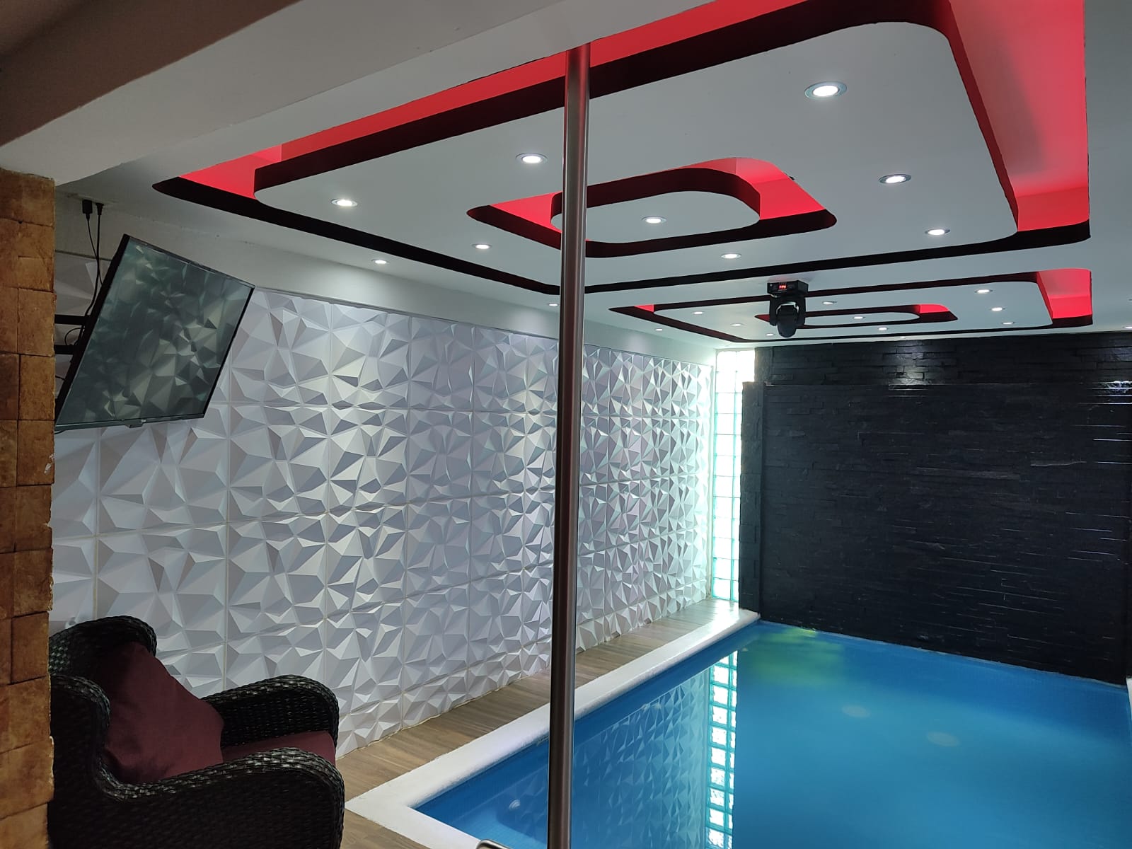 Habitación moderna con piscina interior, televisores montados en la pared, iluminación LED en techo rojo y decoraciones geométricas.