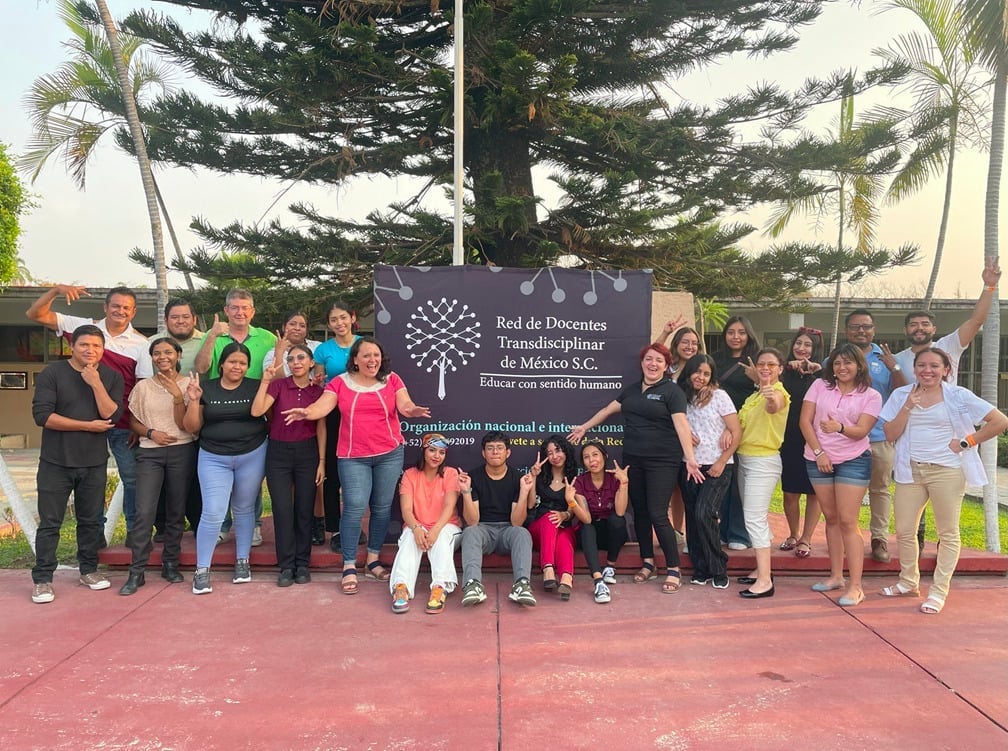 Grupo de personas de pie frente a un cartel de 'Red de Docentes Transdisciplinar de México S.C.' con sonrisas, árboles y cielo despejado.