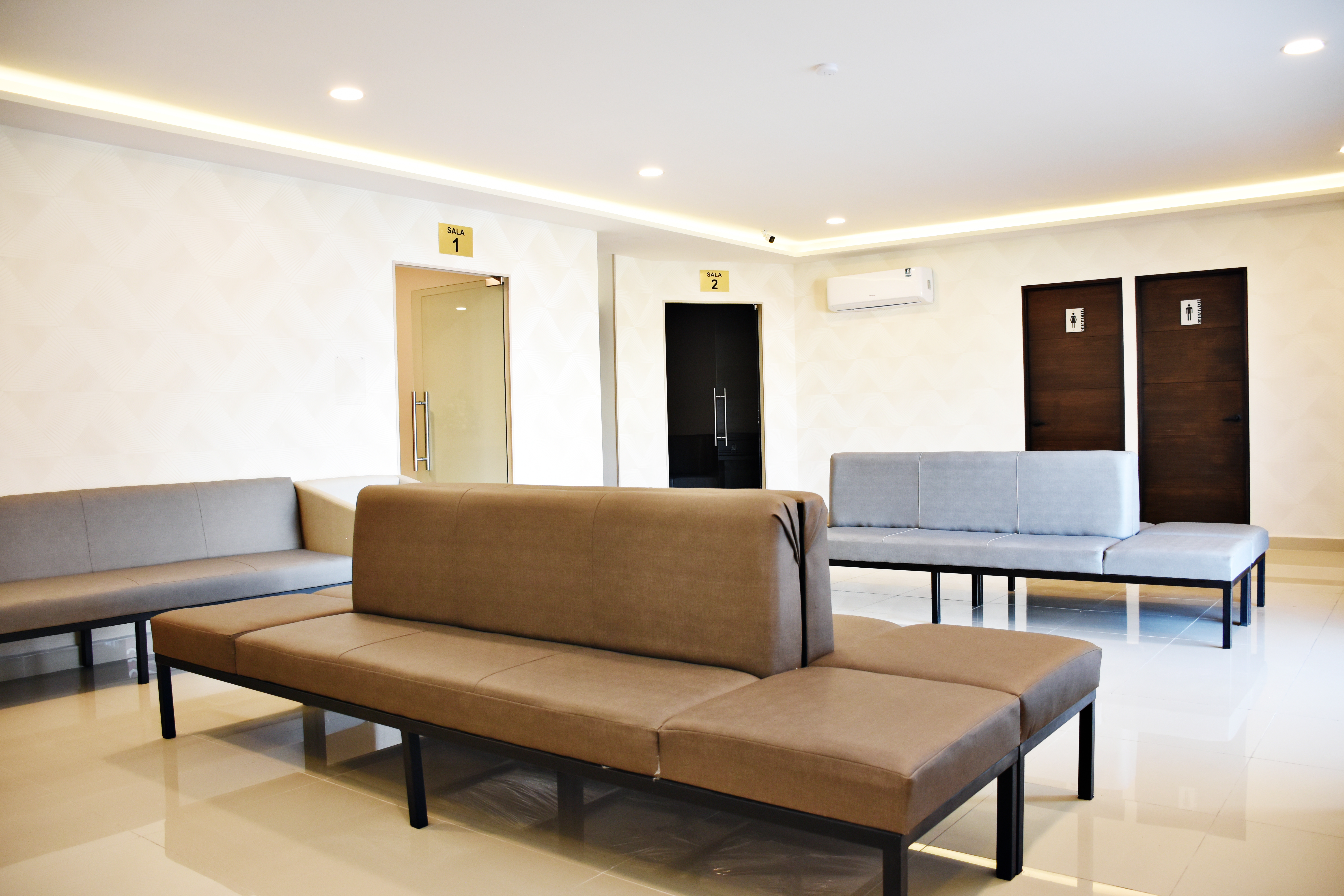 Modern waiting room with upholstered benches, recessed ceiling lights, and doors labeled Sala 1 and Sala 2