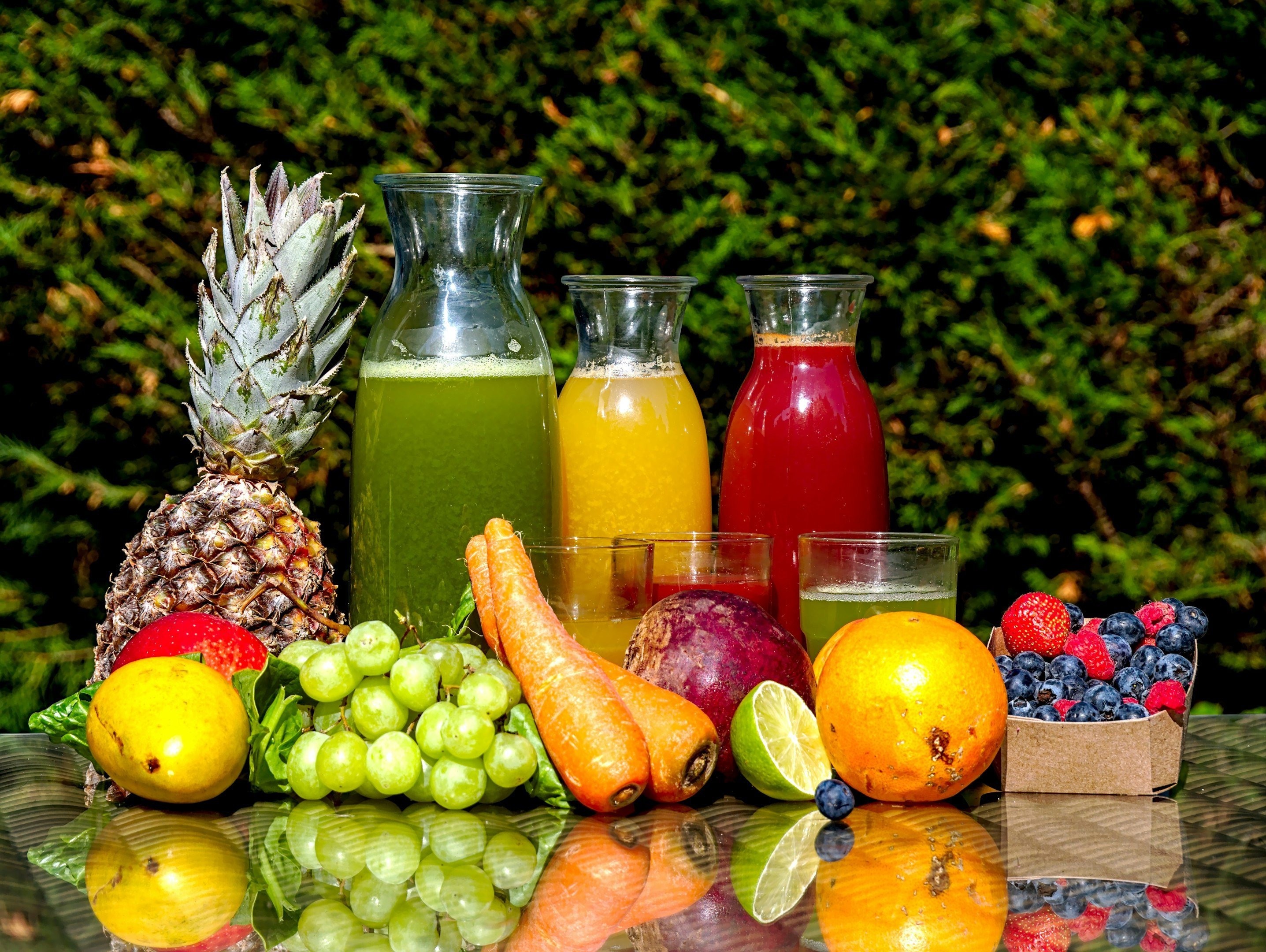 Imagen de una variedad de frutas frescas y jugos en botellas, incluyendo piña, zanahorias, naranja, mango, uvas, y berries, sobre una mesa con fondo natural verde.