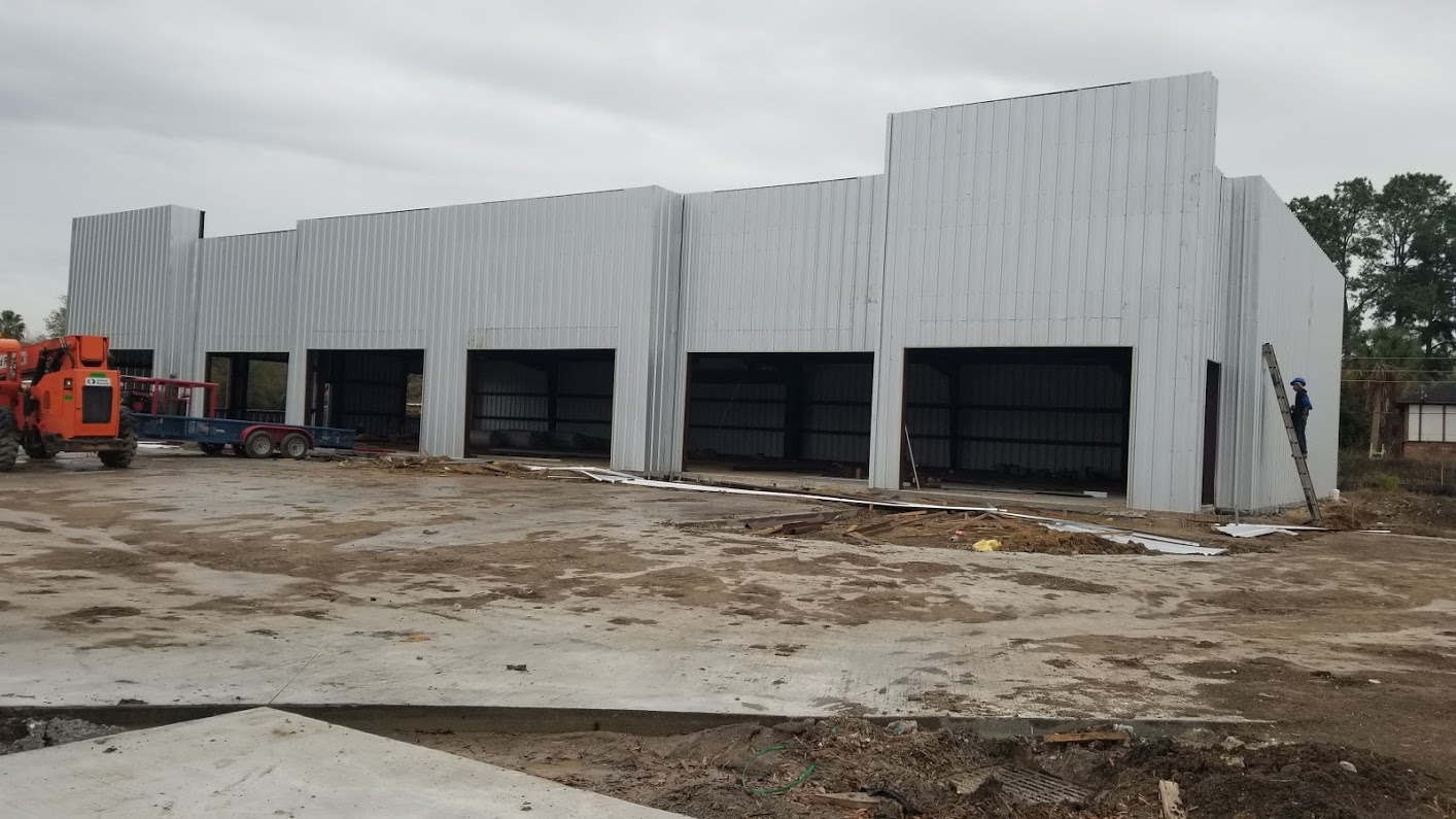 Large industrial building under construction with metal siding, open garage doors, construction equipment, and a worker on a ladder.