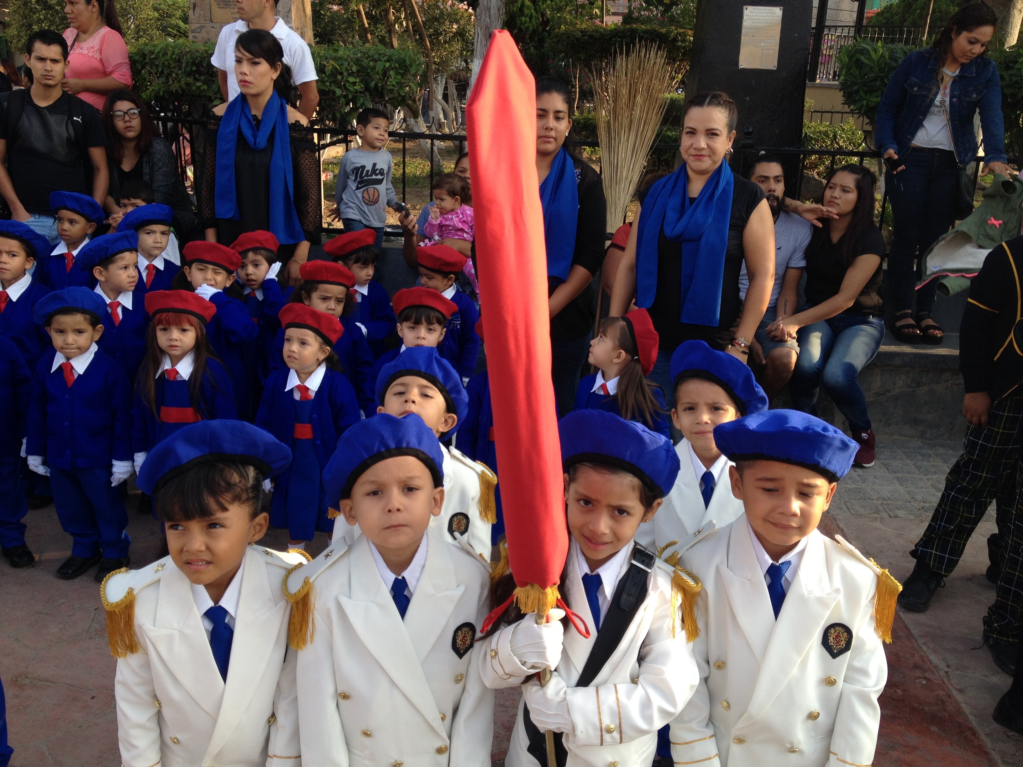 Niños con uniformes blancos y azules en un desfile escolar, sosteniendo una bandera roja, rodeados de adultos y otros niños en un ambiente urbano.