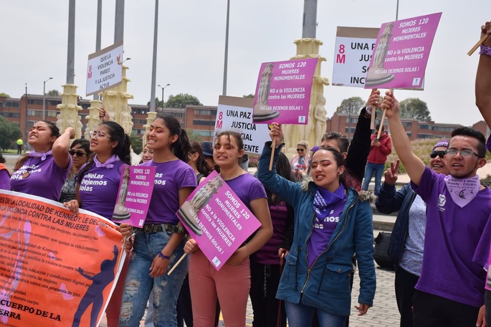 Manifestación de personas con camisetas moradas y carteles protestando contra la violencia hacia las mujeres en un espacio público.