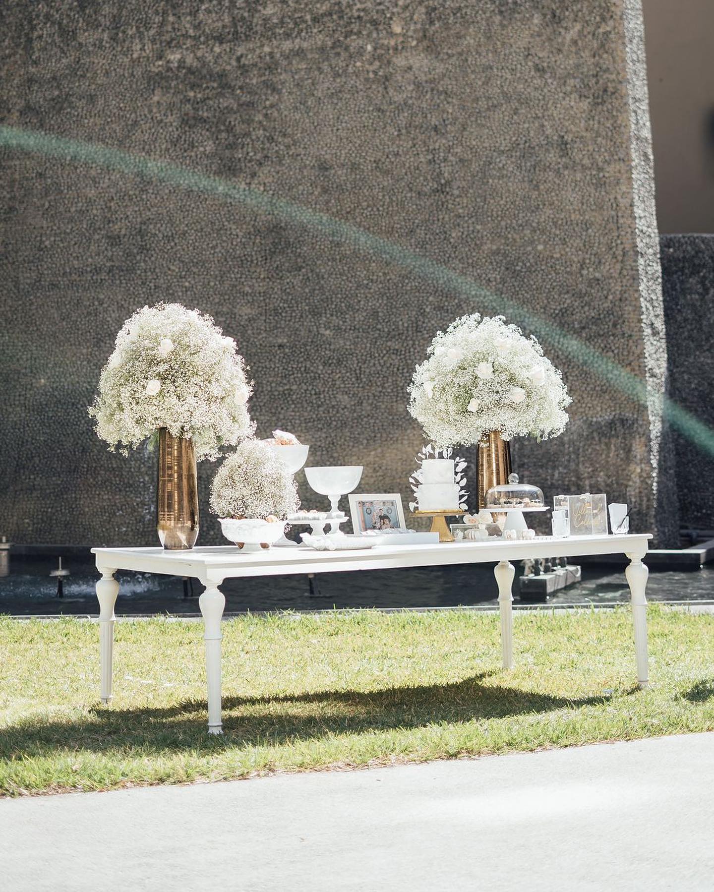 Ramo de flores con rosas rojas, rosadas y melocotón, rodeadas de pequeñas flores blancas y lilas, en un jarrón sobre una mesa junto a una ventana.