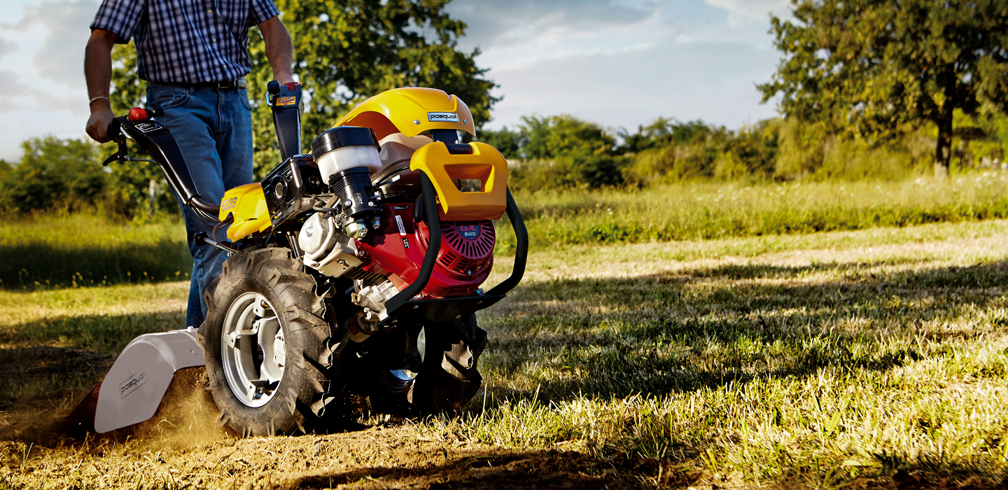 Tractor verde en un almacén con remolque amarillo y vegetación en primer plano.