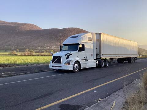 Camión tráiler blanco circulando por carretera rural junto a campos verdes y montañas al atardecer Camión tráiler blanco circulando por carretera rural junto a campos verdes y montañas al atardecer