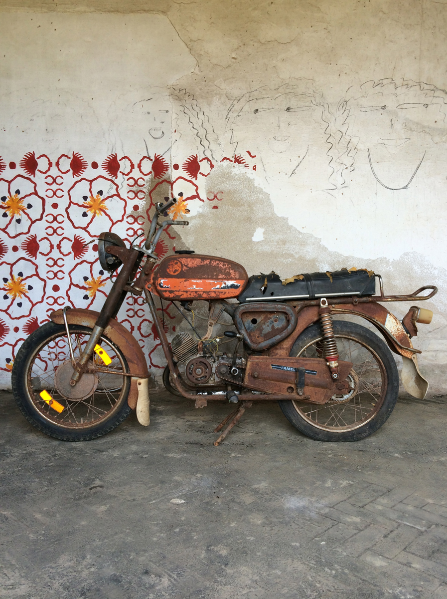 A rusty motorcycle against a wall with a floral mural and three hand-drawn faces.
