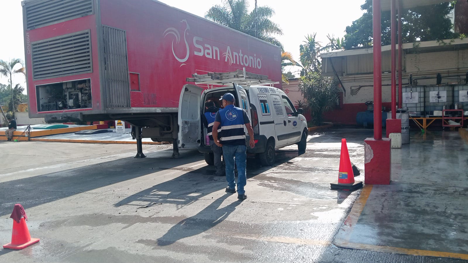 Camioneta de servicio técnico al lado de un camión rojo en un estacionamiento, con dos trabajadores revisando equipos, rodeados de señales de seguridad y vegetación.