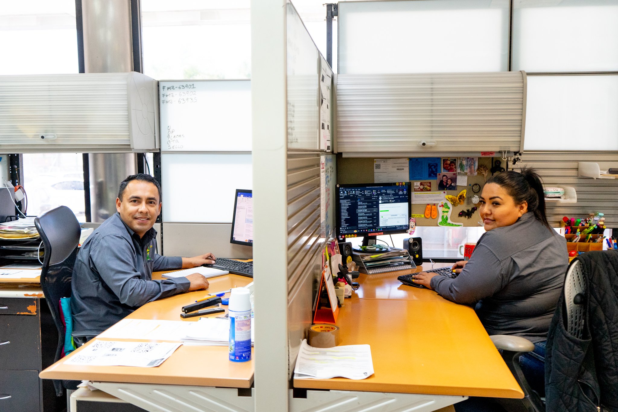 Dos personas trabajando en cubículos de oficina con computadoras y papeles sobre el escritorio.