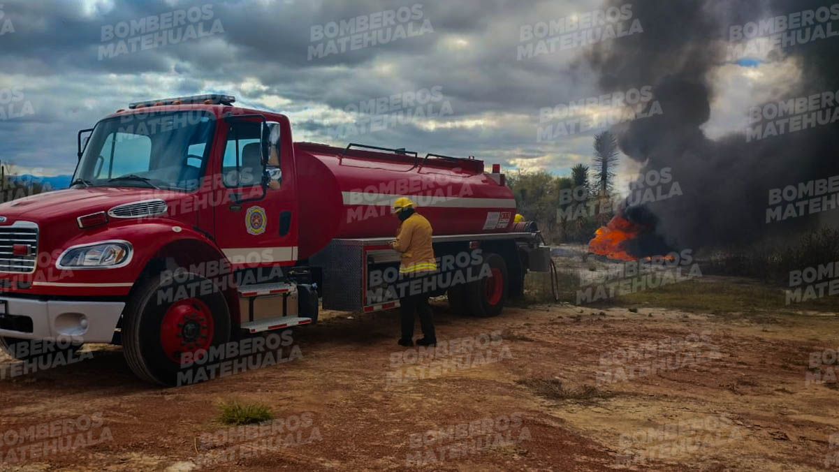 Bomberos con equipo de protección frente a un camión de bomberos, con un gran incendio al fondo en la ciudad, de noche.