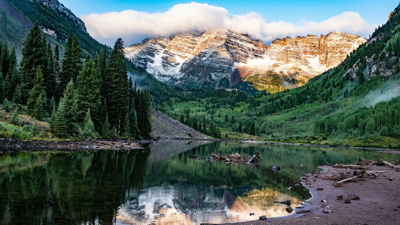 Scenic view of Maroon Bells mountains in Colorado with snow-capped peaks, pine trees, and a reflective lake.
